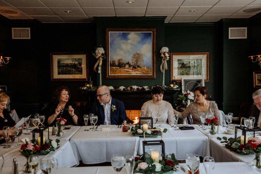 A bride and groom are sitting and talking to family members around a large table in front of a dark green wall decorated with flowers and photos during their wedding reception
