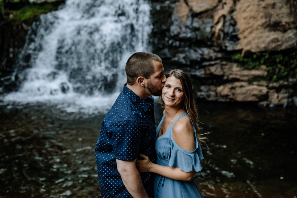 Girl looking at camera while boy kisses her on the cheek with waterfall in the background