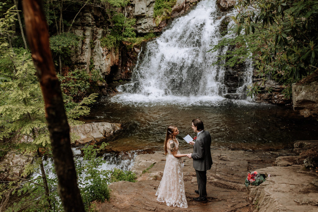 Man reads from a vow book while holding a womans hand during an intimate wedding ceremony in front of a waterfall at Hickory Run State Park in Pennsylvania