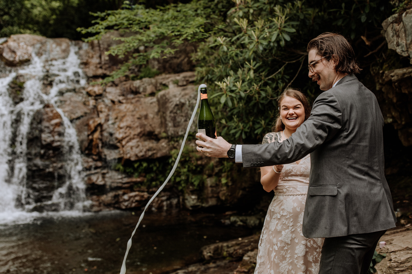 Man and woman popping champagne in front of a waterfall after wedding ceremony