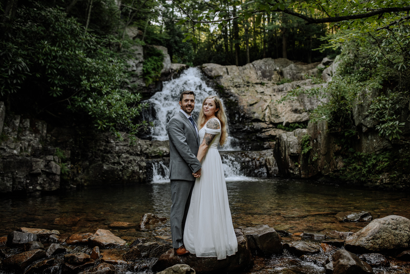 Man and woman posing (looking at camera) in wedding attire kissing in front of Hawk Falls in Hickory Run State Park