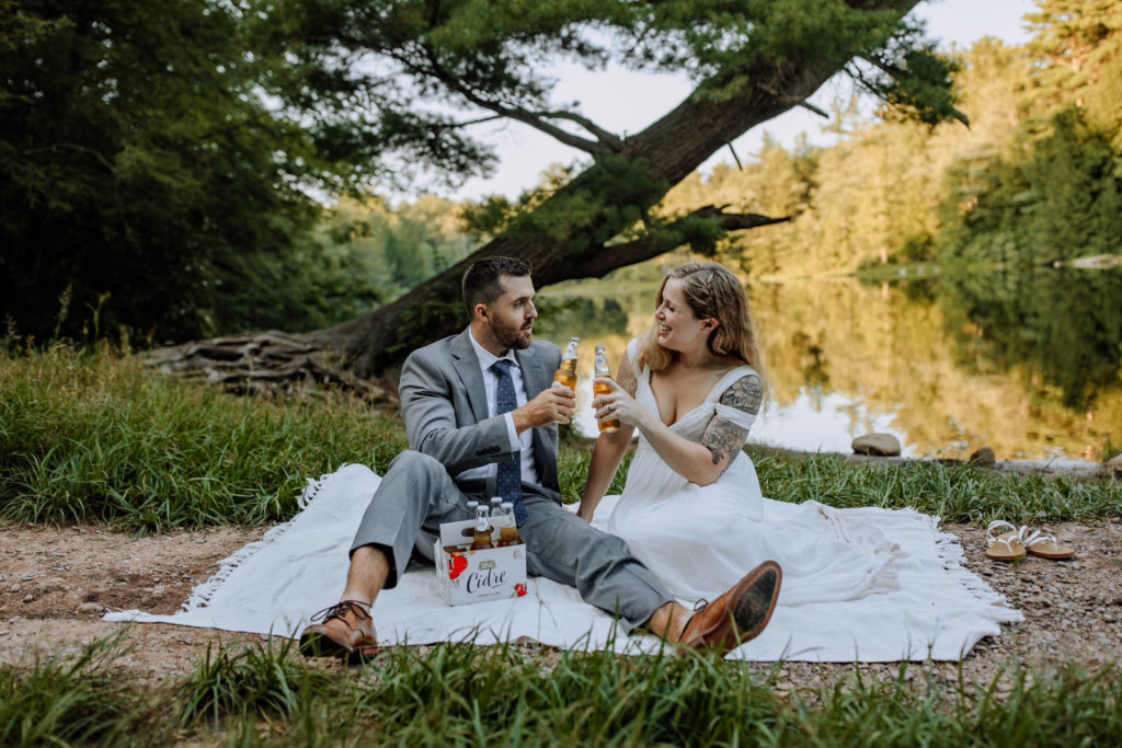 Man and woman sitting on a blanket drinking hard apple cider in front of a lake