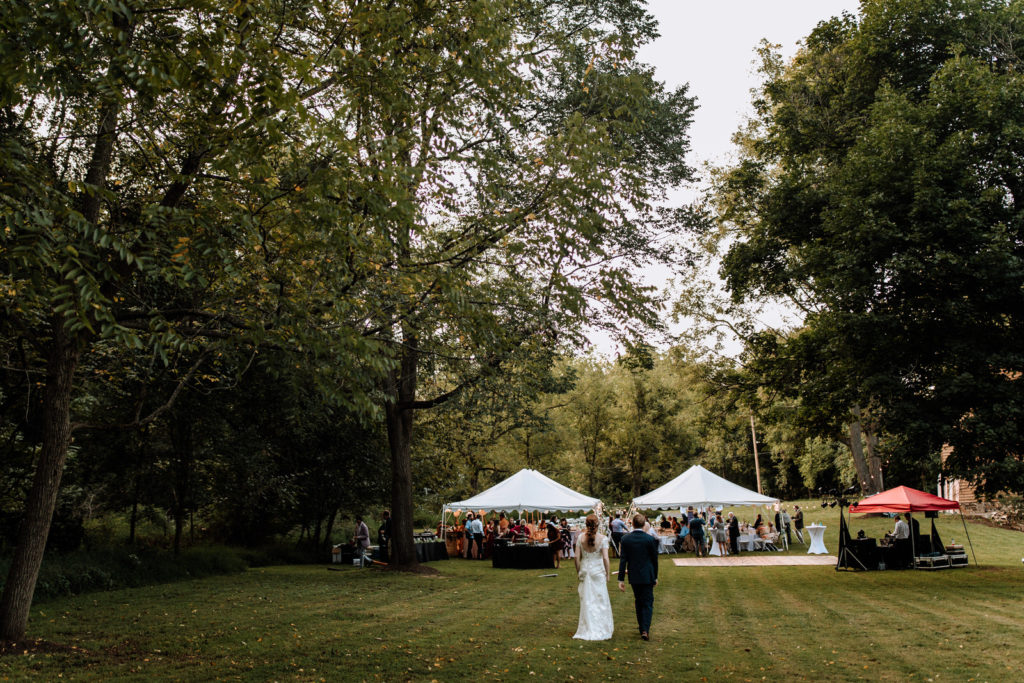 A man and woman walk towards 2 large tents for their outdoor wedding reception in the woods