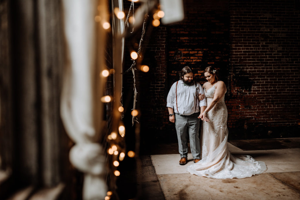 Man and woman dressed in wedding garb standing in an industrial brick building with string lights in the foreground