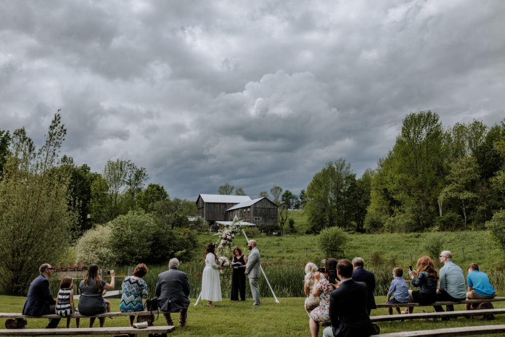 A wide view of a small wedding ceremony in front of a lake and barn