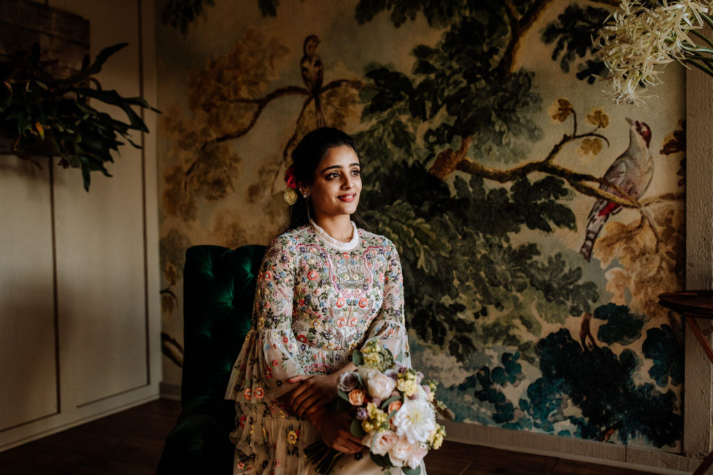An Indian woman dressed in a floral wedding gown is sitting on a green chair in front of a decorative wall for a bridal portrait with a bouquet of flowers in her hands