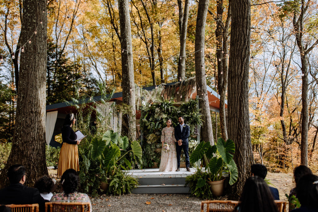 Indian bride and groom standing on wooden altar in front of a wall of green plants while a marriage ceremony is officiated at Promise Ridge in the Poconos, PA.