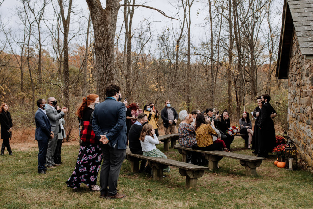 Family and friends view a bride and groom dressed in black attire get married in front of a stone building at Jacobsburg Park in PA