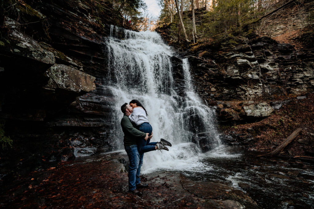 A man holds a woman up in front of a large waterfall and kisses her