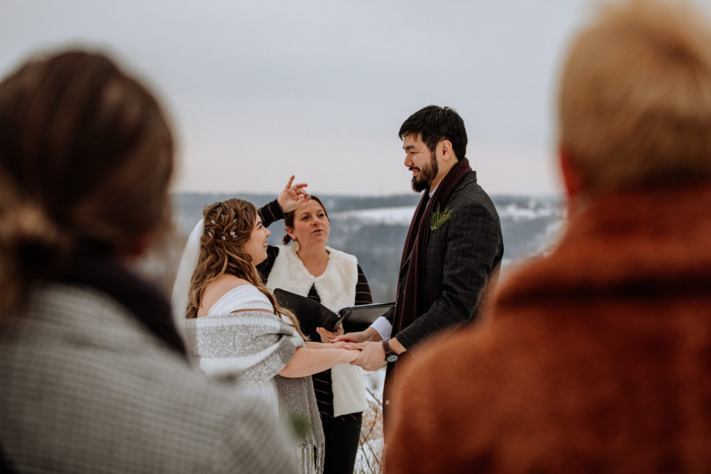 A wedding officiant holds a wedding ring in front of a bride & groom who are holding hands during a small wedding ceremony as family watches