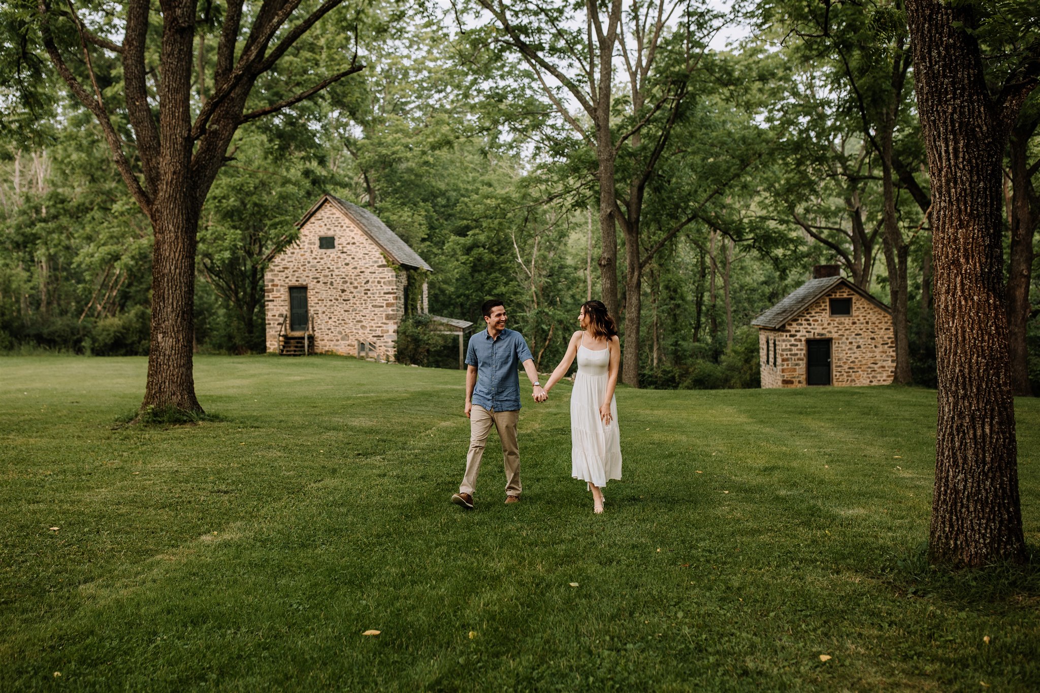 Engagement photo session as Jacobsburg Park with couple walking holding hands and two rustic houses in the background