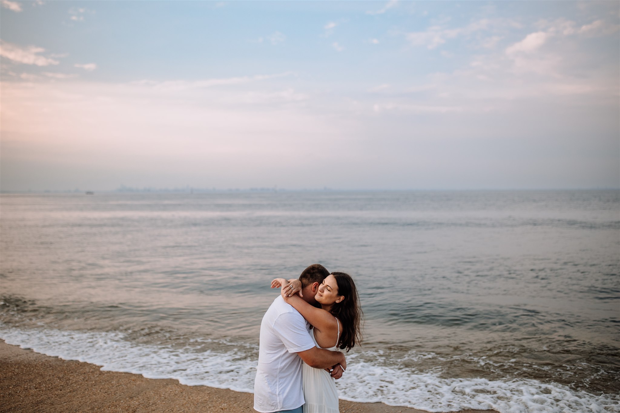 Couple hugging on beach at Sandy Hook in New Jersey with sunset behind them