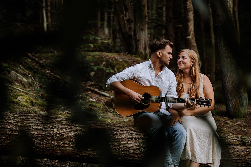 Man dressed in white shirt and jeans playing guitar to a woman in a sun dress while sitting on a log