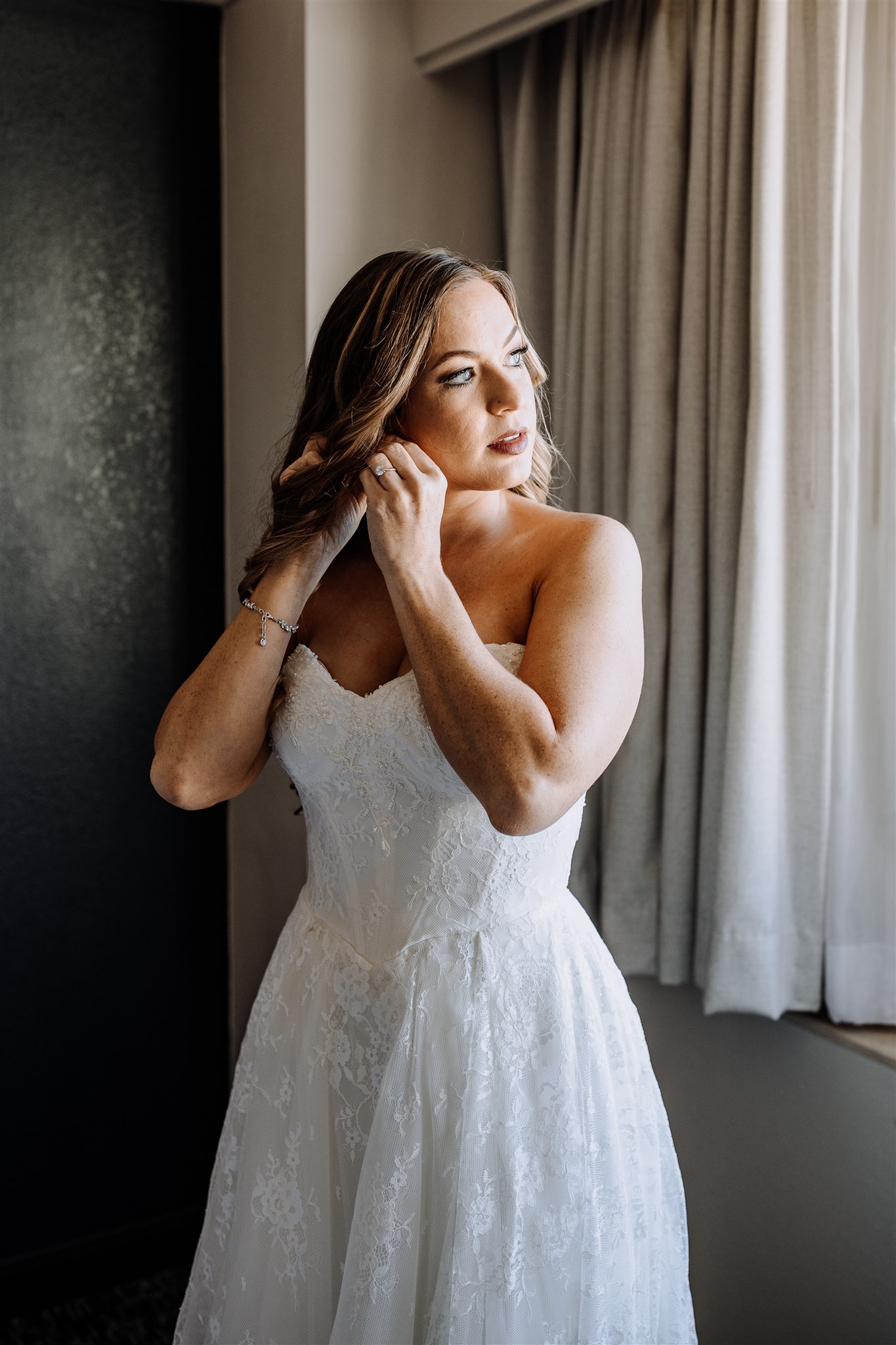 Bride in wedding dress looking out window while fixing earing